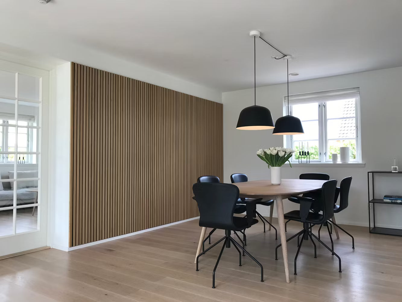 Modernes Esszimmer mit Holzboden, Akupanel-Akustiklattenwand, schwarzen Stühlen, Holztisch, Pendelleuchten und weißem Blumenschmuck.