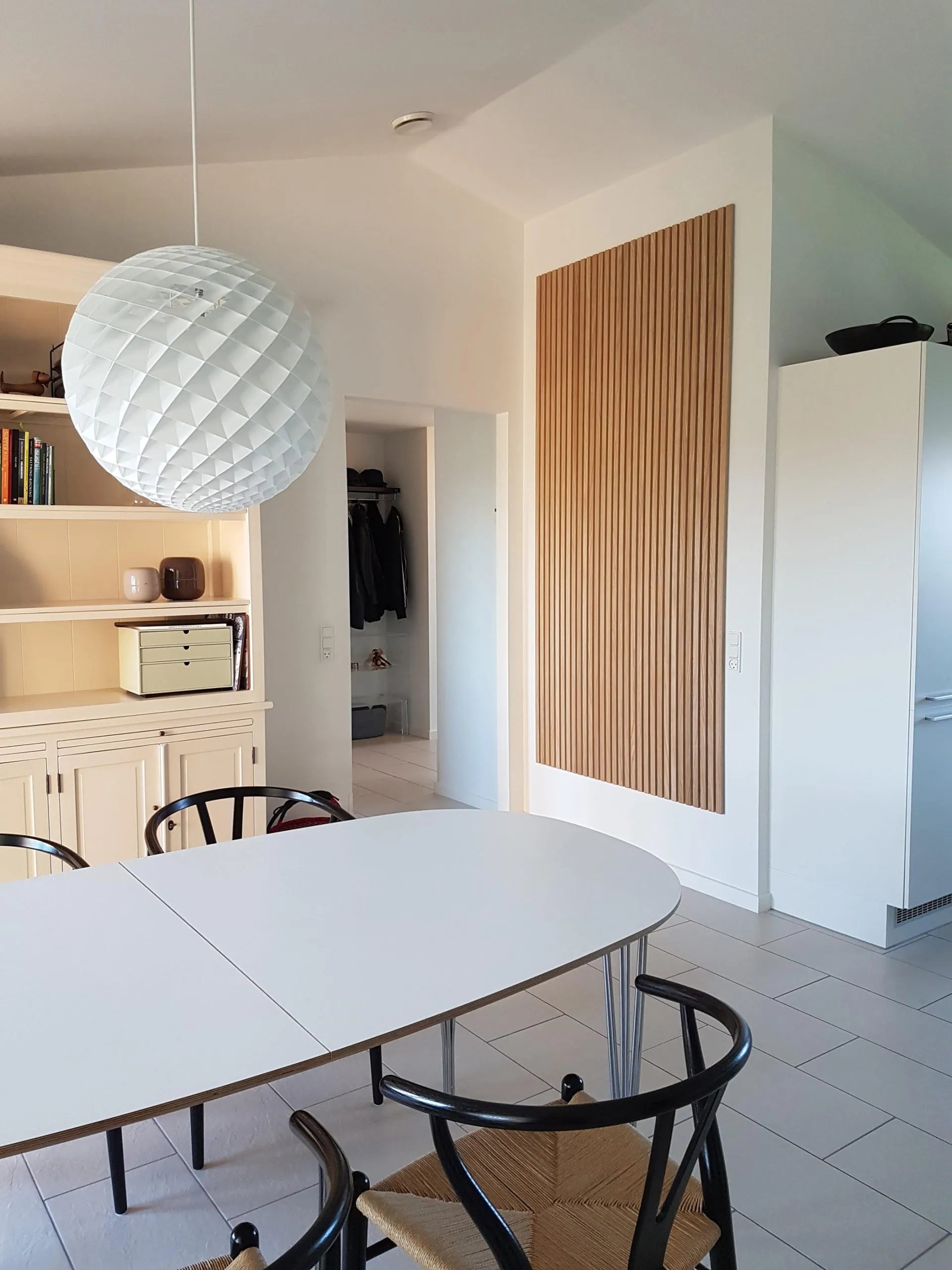 Moderner Essbereich mit einem weißen ovalen Tisch, schwarzen Stühlen, einer großen strukturierten Hängeleuchte und eleganten Akupanel-Akustikschränken aus hellem Holz.