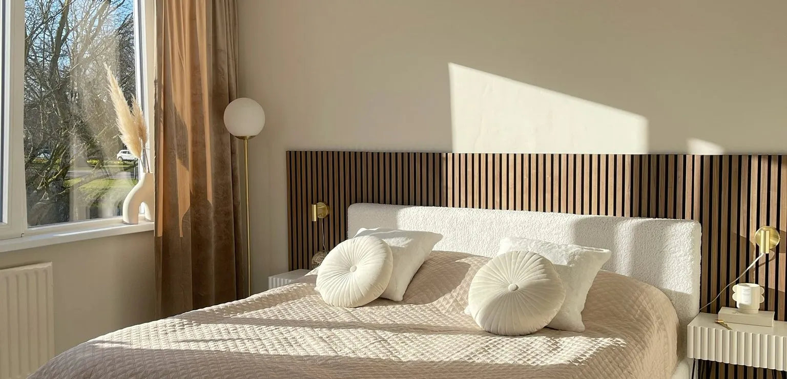 Sonnendurchflutetes, modernes Schlafzimmer mit einem beigen Steppbett, runden Kissen, einem Kopfteil aus Akupanel-Holzlatten und minimalem Dekor neben einem großen Fenster mit transparenten Vorhängen.