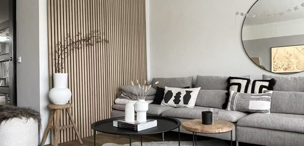 Modernes Wohnzimmer mit einem grauen Sofa, abstrakten Kissen, schwarzen Couchtischen, dekorativen Vasen und einer Akupanel-Holzlattenwand mit rundem Spiegel.