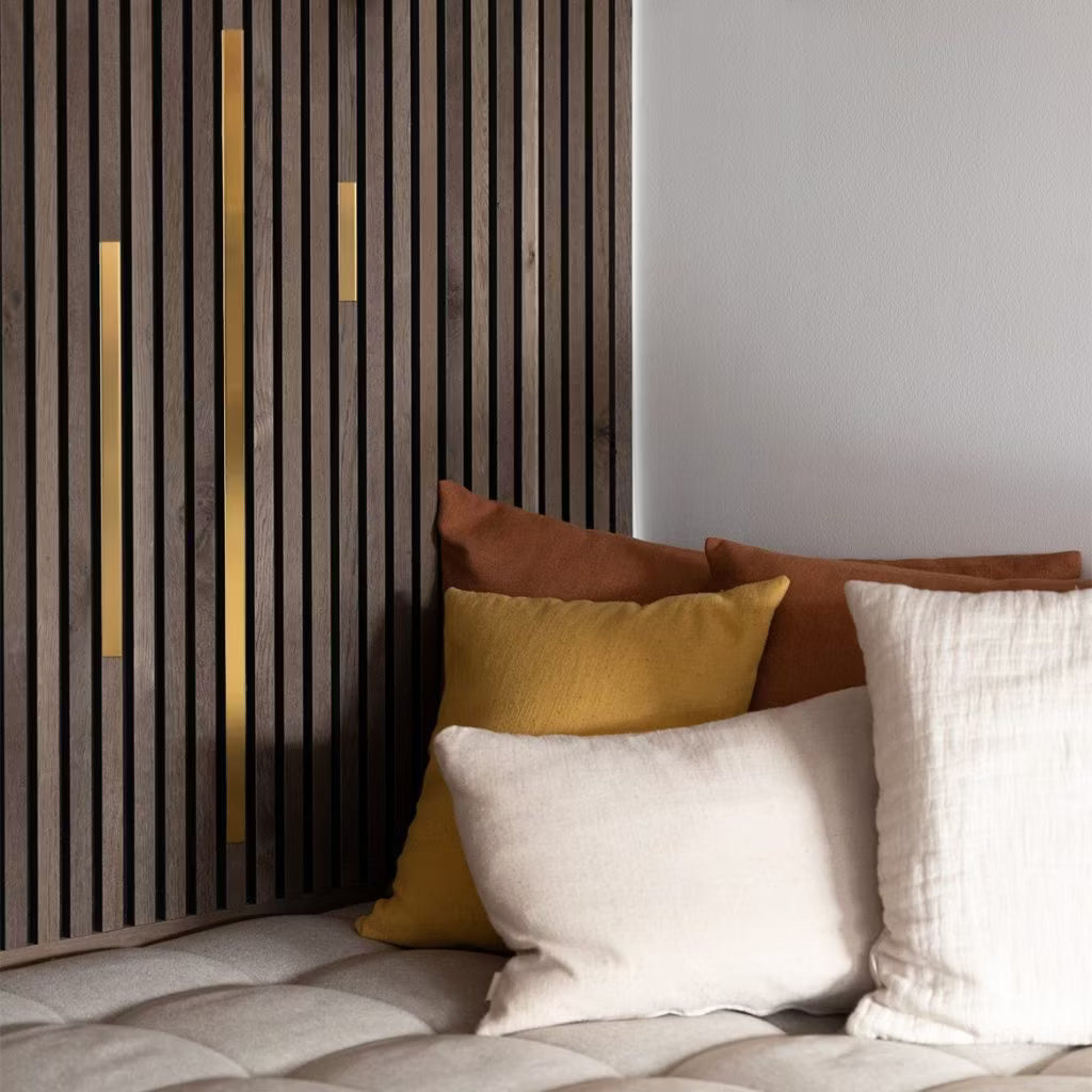 Cremefarbene, gelbe und braune Kissen auf einem beigen, getufteten Sofa neben einer modernen Akustik-Lamellenwand mit goldenen Akzenten sorgen für ein gemütliches, modernes Ambiente.