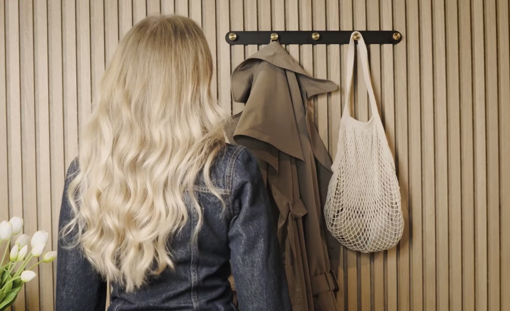 Eine Frau mit langen blonden Haaren steht vor einem Kleiderständer mit einem Trenchcoat, einer weißen Netztasche und frischen weißen Tulpen neben einer Akupanel-Holzplatte in der Ecke.