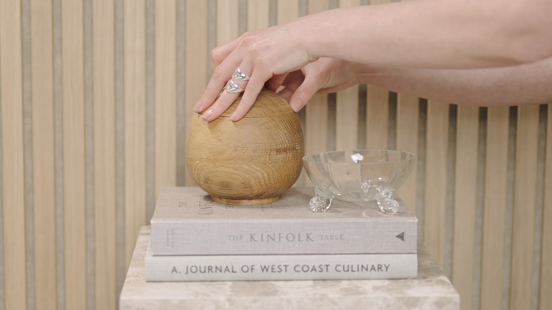 Eine Hand berührt eine Holzkugel, die auf gestapelten Büchern, darunter Kinfolk Table und Journal of West Coast Culinary, neben einer Glasschale auf einer Akustikplatte aus Holzlatten steht.