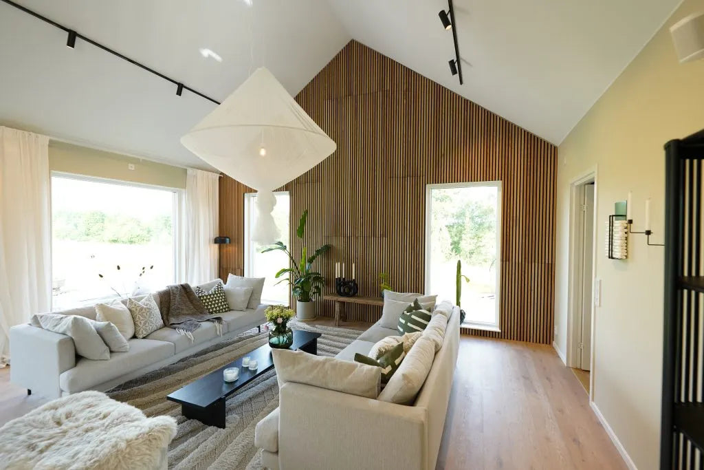 Modernes Wohnzimmer mit hoher Decke, großen Fenstern, hellen Sofas, Akupanel-Akustikholz-Akzentwand, Zimmerpflanzen und viel Tageslicht.