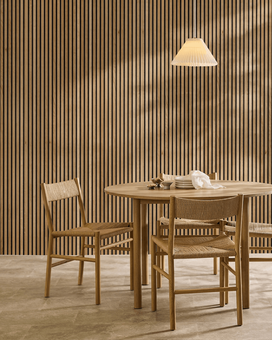 Minimalistischer Essbereich mit einem Holztisch, zwei Stühlen und einem WoodUpp Akupanel Akustik-Holzpaneel in Rustic Brown Oak (300x60 cm), beleuchtet von einer Hängelampe.