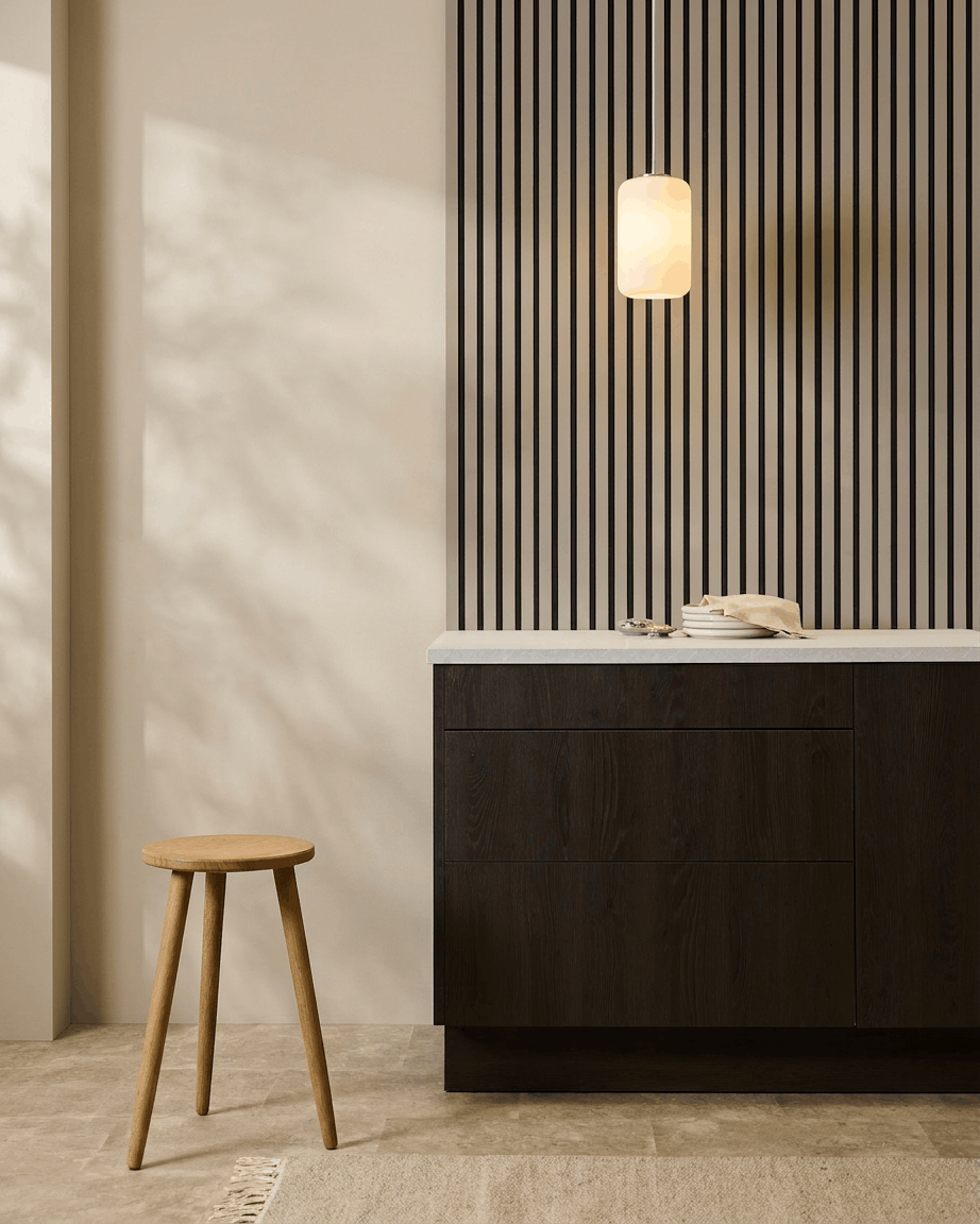 Minimalistische Küche mit WoodUpp Akupanel (Stone, Black Felt) Akustikpaneelen an der Wand, Holzstuhl, dunklen Schränken und Pendelleuchte in neutralen Tönen.
