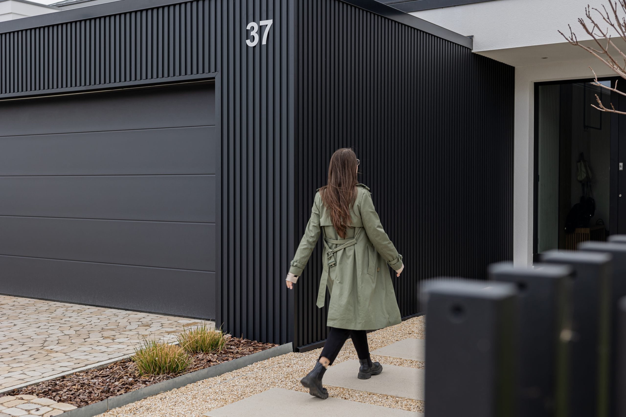 A woman in a green coat walks toward the entrance of modern house 37, featuring a sleek black Aluwood facade panel that highlights contemporary architecture.
