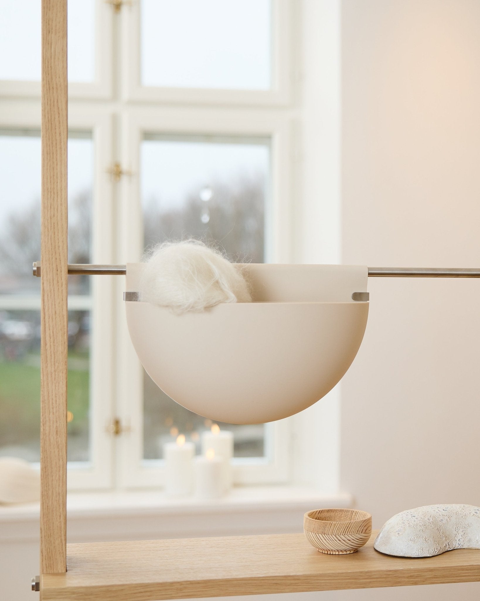 Eine weiße Schale mit einem flauschigen Objekt steht auf einem Holzregal neben Kerzen, Deko und einem WoodUpp Gro Soft Pocket BM2 Akustik-Accessoire in hellem beige am Fenster.