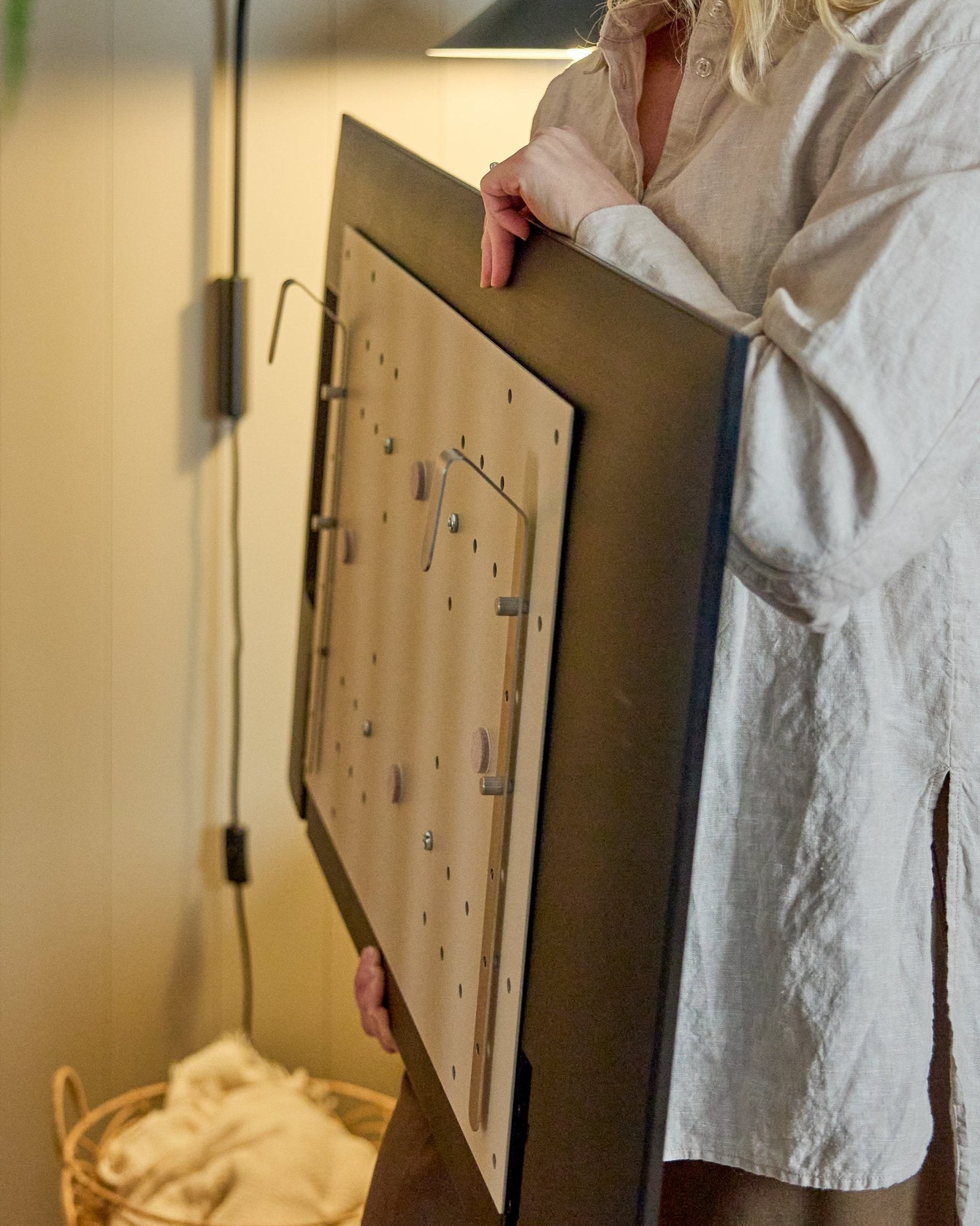 Eine Person in einem hellen Hemd hält die WoodUpp Nell BM1 TV-Mount in beige, 64x44 cm – kompatibel mit nordischen Akustik-Holzpaneelen, für Installationen ohne Wand.