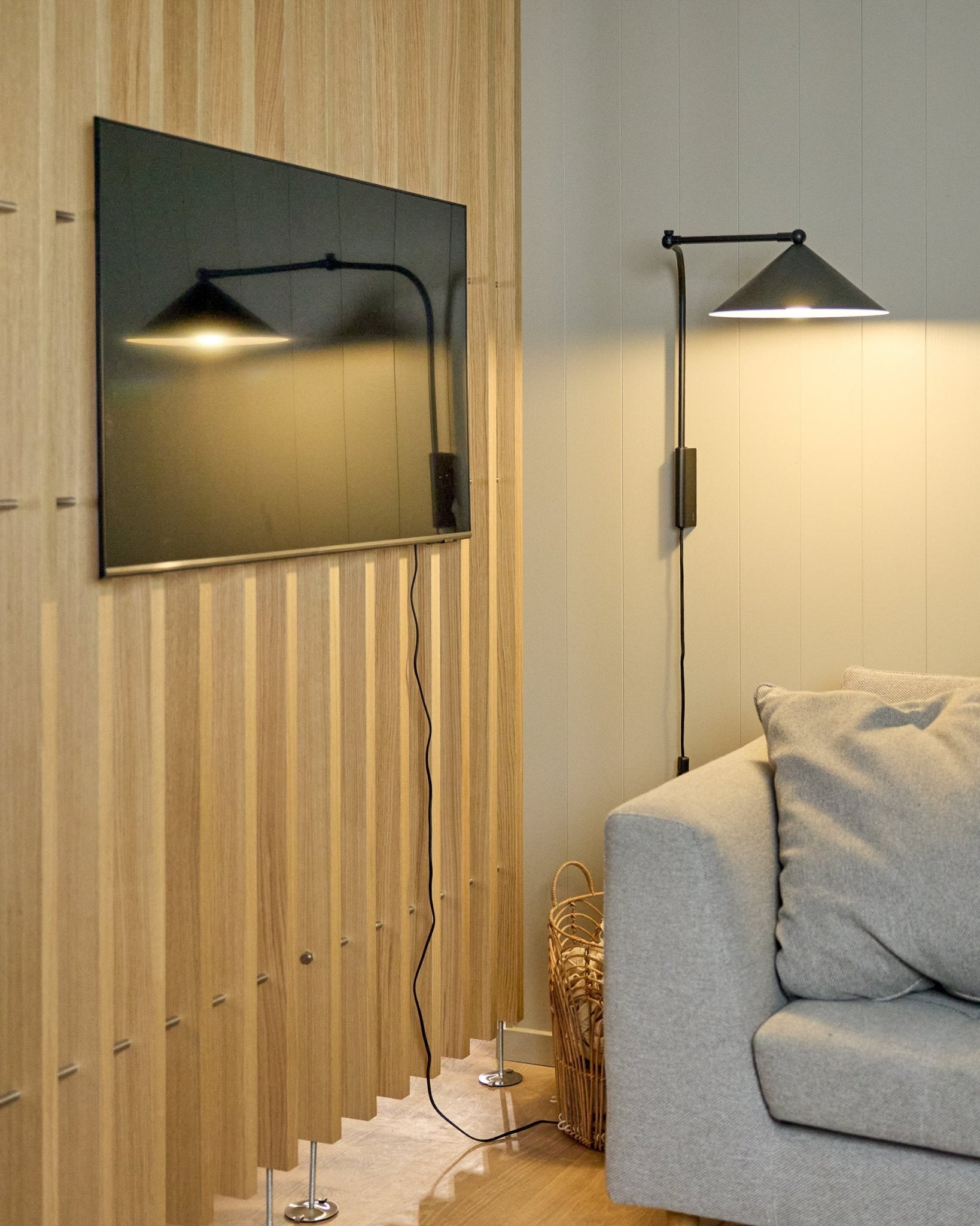 Ein Nell | TV-Mount / BM1 von WoodUpp (Beige, 64x44 cm) befestigt einen Flachbildfernseher an einem nordischen Akustikpaneel neben einem grauen Sofa; ein Korb steht in der Nähe.