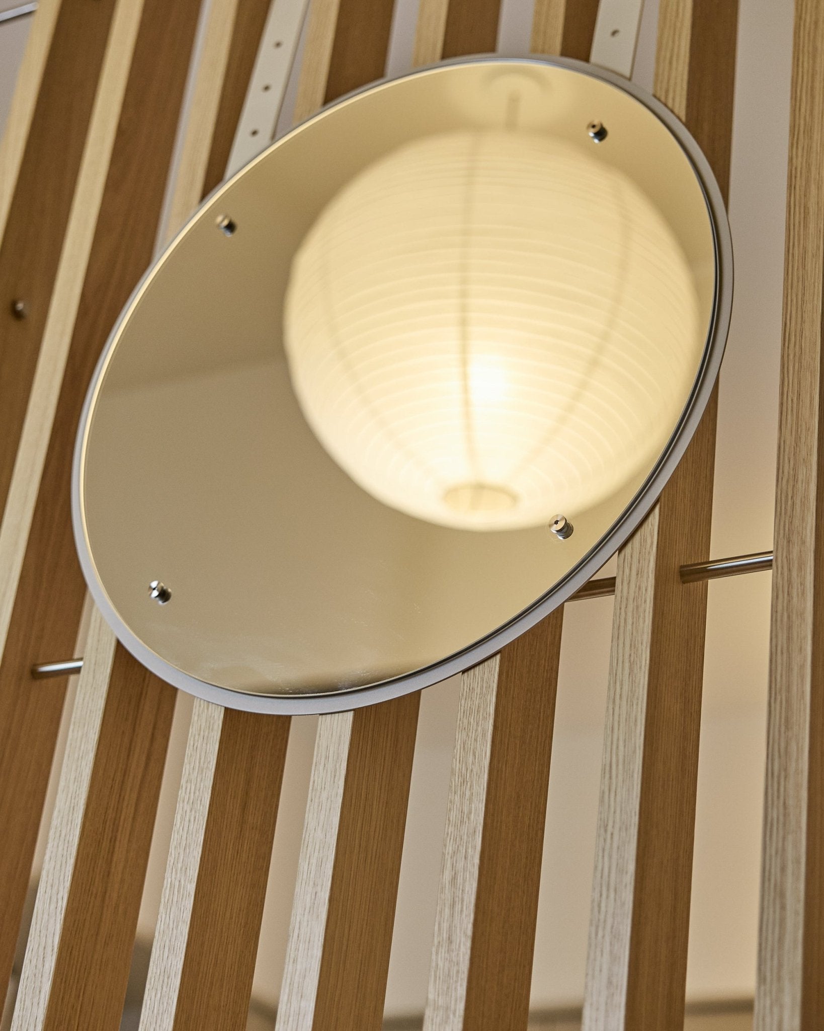Der Vala | Mirror / BM1, D54 cm von WoodUpp ist ein höhenverstellbarer runder Spiegel, der auf vertical Akustik-Lamellenpaneelen montiert ist und eine leuchtende Papierlaterne reflektiert.