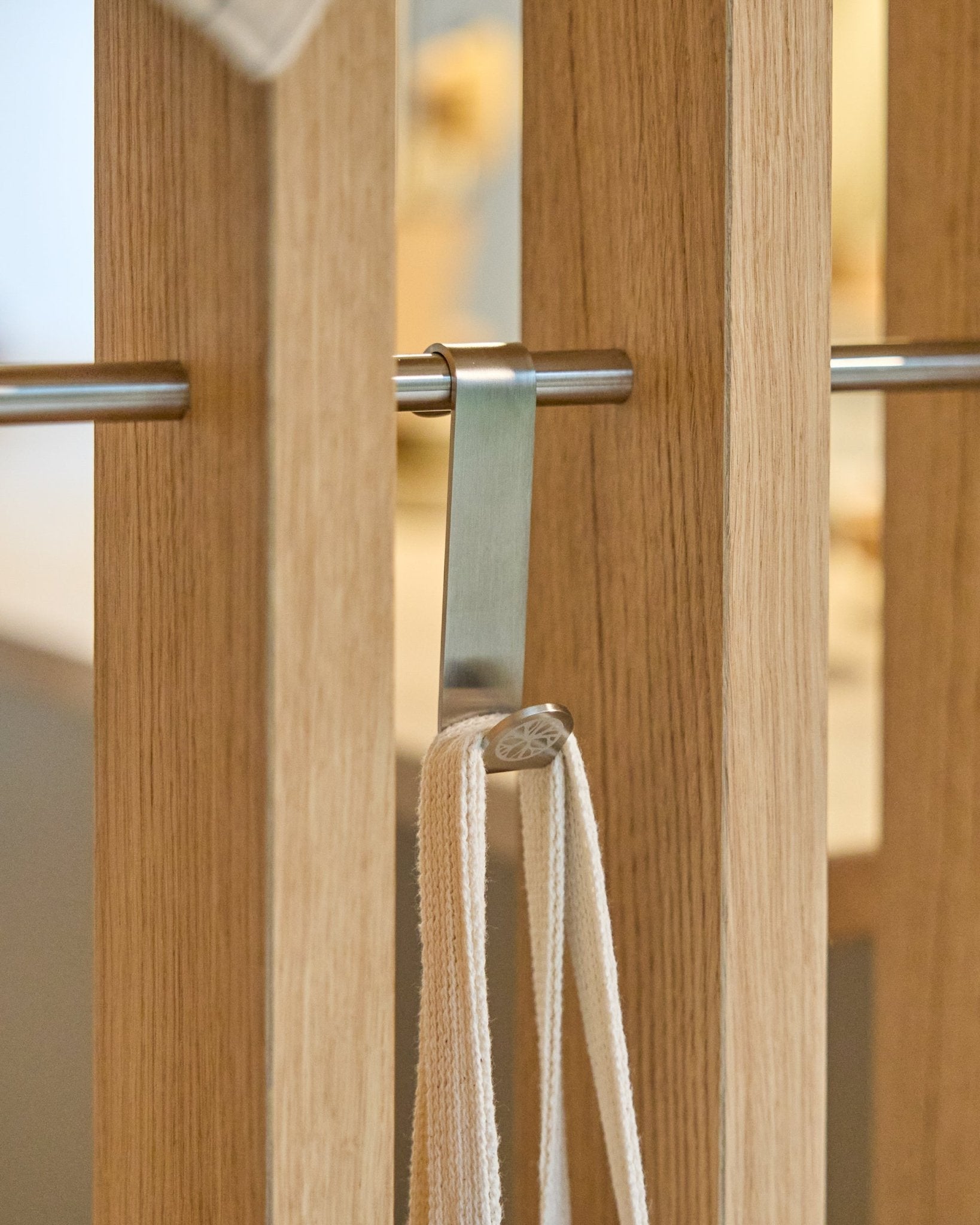 Ein WoodUpp Alva S-Haken aus steel Edelstahl befestigt eine Schlaufe mit beige an einer Stange, die zwischen vertical Akustikpaneelen mit Holzlamellen montiert ist. Das Set enthält 2 Haken.