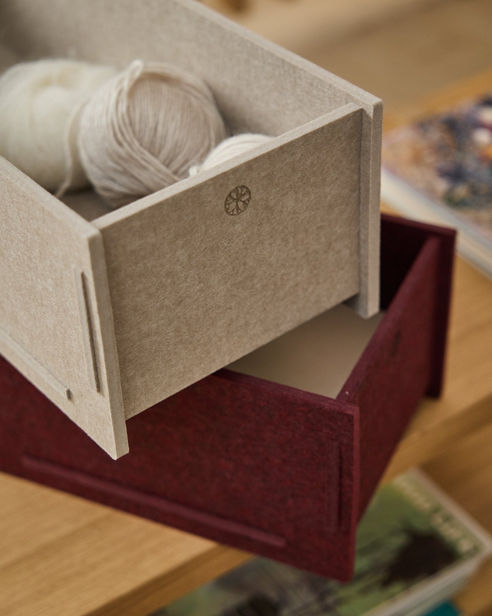 Eine beige PET-Filz-Aufbewahrungsbox, gefüllt mit Garn, steht auf einer geschlossenen WoodUpp Vigg Felt Box/BM2 in ox red auf einem Holzboden, mit Büchern im Hintergrund.