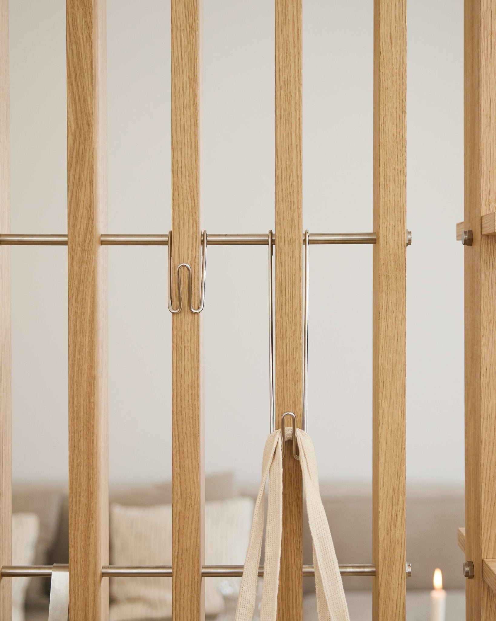Nahaufnahme des WoodUpp Alva | Wire Hook BM1 (Edelstahl steel, lang, 3er-Set), befestigt an hellen Akustikpaneel Lamellen mit einem weißen Band; Sofa und Kerze sind im Hintergrund unscharf.
