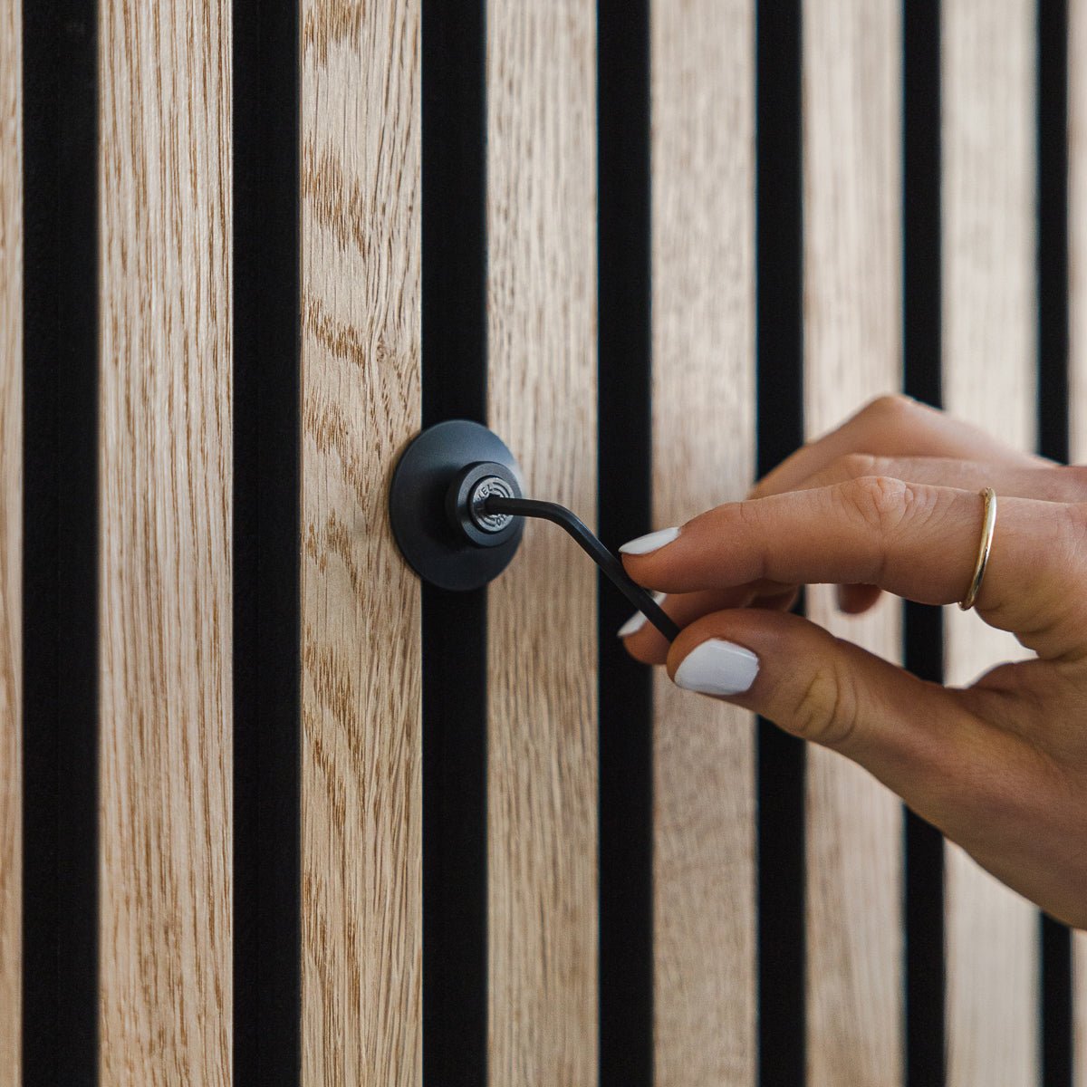 Eine Hand mit weißem Nagellack dreht einen Dupps | Heavy black Aluminiumhaken von WoodUpp an einer vertical Akupanel Akustik-Holzplatte.
