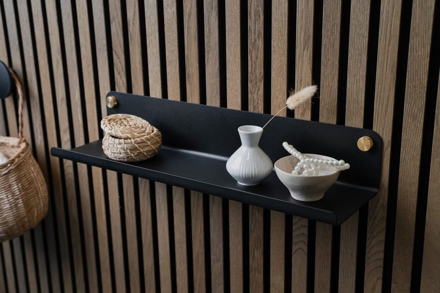 Ein schwarzes Wandregal mit einem kleinen Weidenkorb, einer weißen Vase und einer Keramikschüssel mit Perlen, montiert auf einer nordischen Akustik-Lamellenwand.